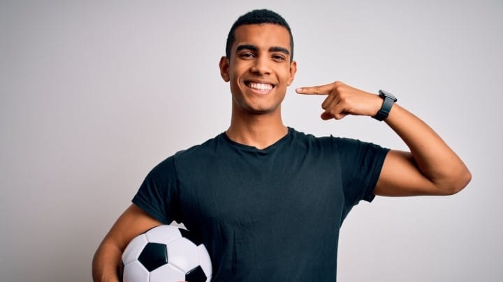 Smiling man holding a soccer ball and pointing at his teeth.